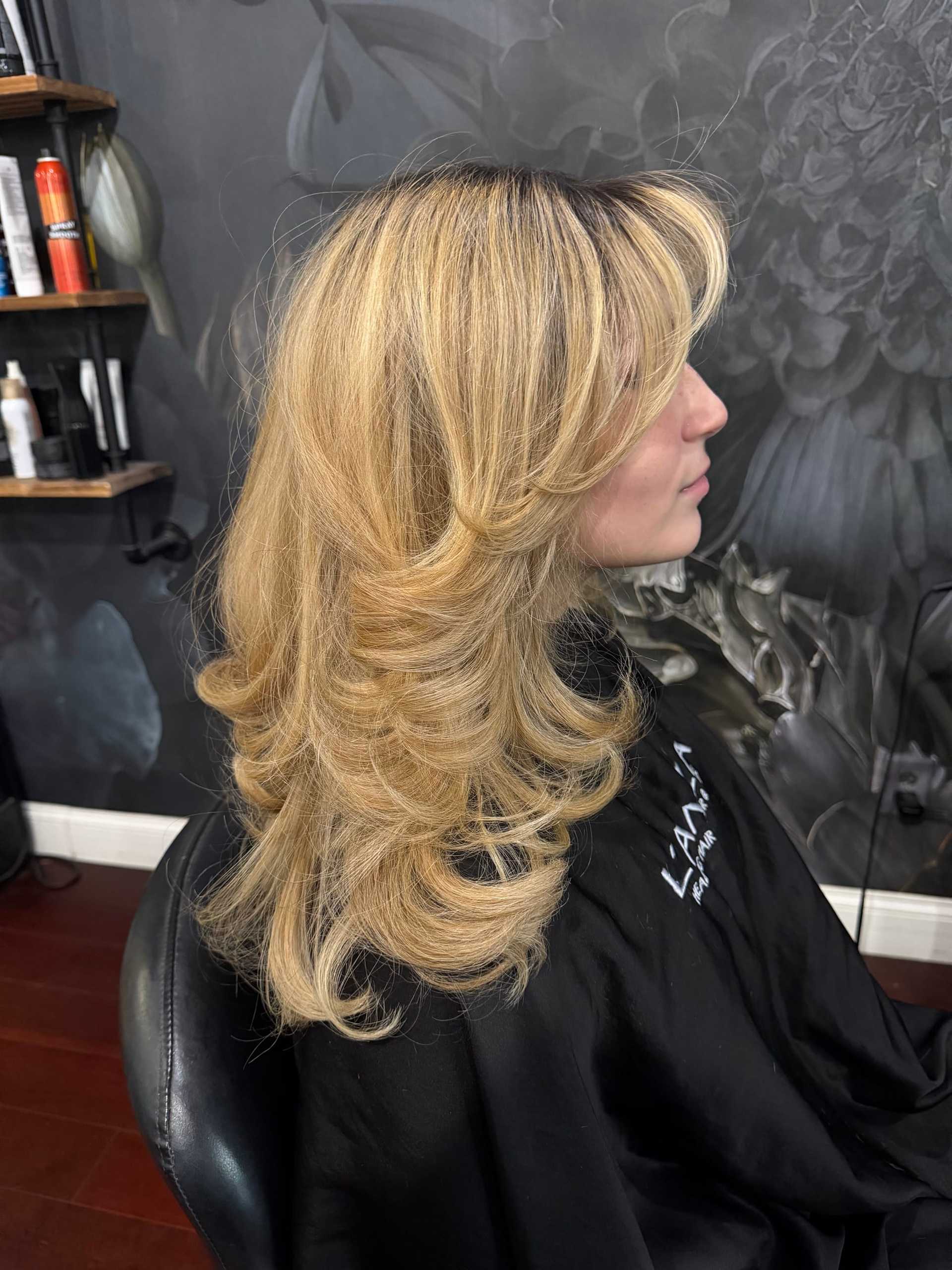Blonde woman with layered hairstyle sitting in a salon chair, gray floral wallpaper background.