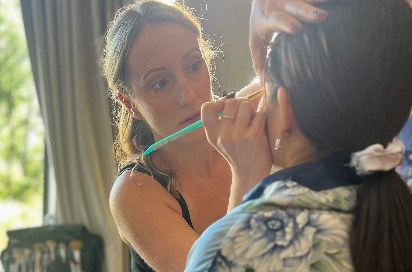 Makeup artist applying makeup to a woman in a well-lit room with a window in the background.