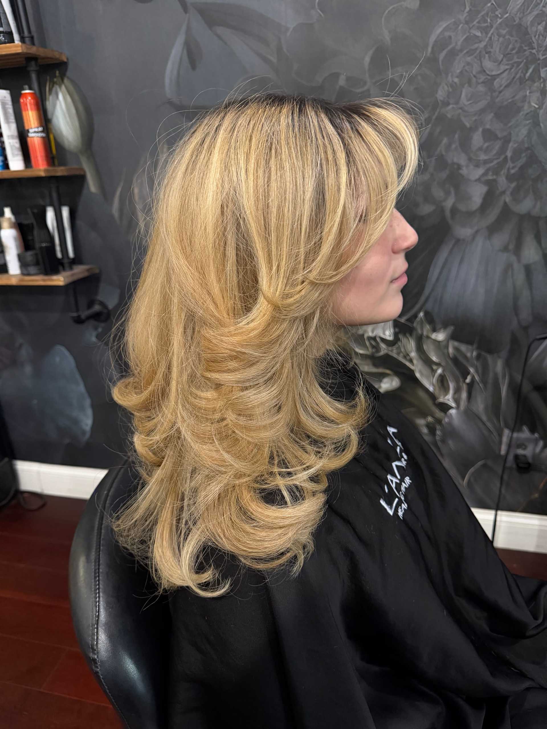 Blonde woman with layered hairstyle sitting in a salon chair, gray floral wallpaper background.